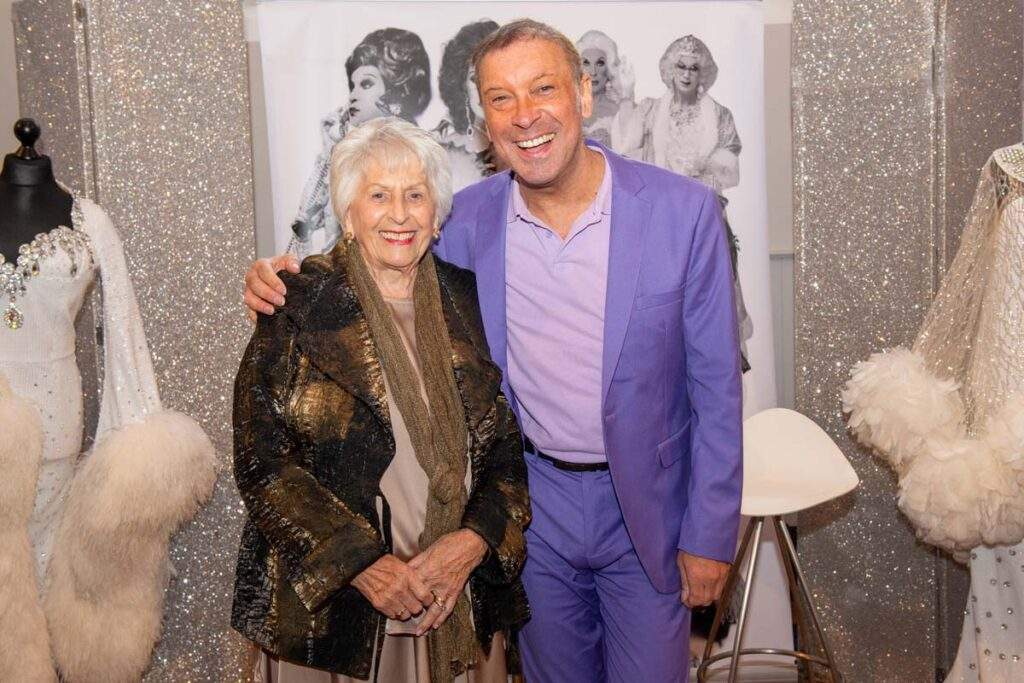 Ceri with one of the female VIP guests in front of an extravagant white feathered costume, with a sparkling silver backdrop. Both are smiling to camera.