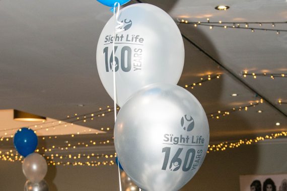 Pic of three balloons - 2 white with the Sight Life logo on them and one blue. They're in a tier as a table centrepiece with a party room in the background.