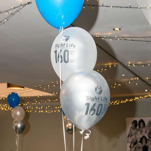 Pic of three balloons - 2 white with the Sight Life logo on them and one blue. They're in a tier as a table centrepiece with a party room in the background.