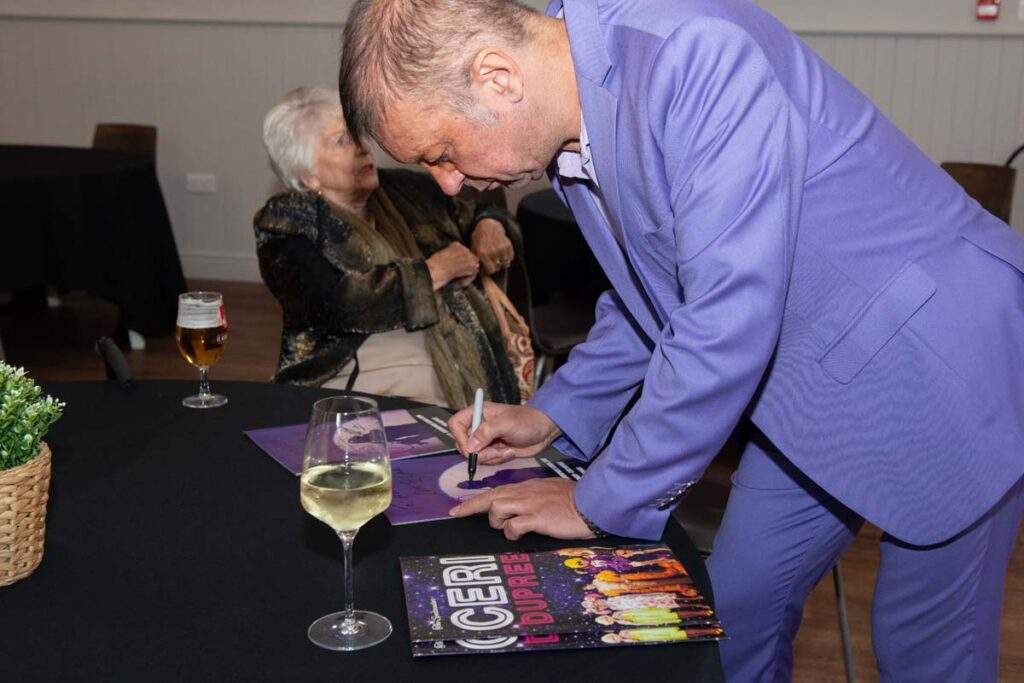 Ceri Dupree leaning over a table to sign a souvenir programme for a guest, dressed in his distinctive lilac suit.