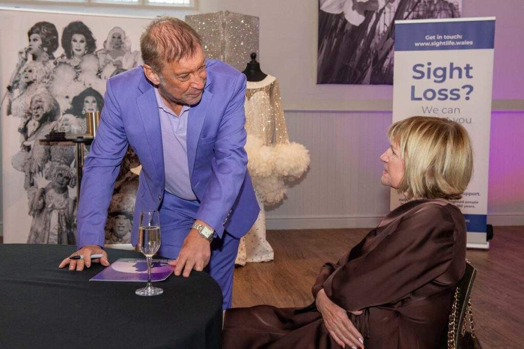 Ceri Dupree chatting warmly with a seated guest, with one of his spectacular white feathered costumes and a Sight Life banner in the background.