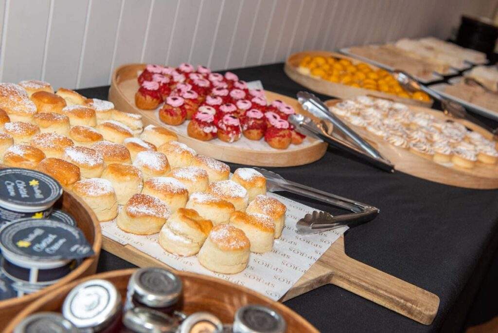 A delicious afternoon tea buffet featuring freshly baked scones, colourful cakes, pastries and cream, beautifully arranged on wooden serving boards.