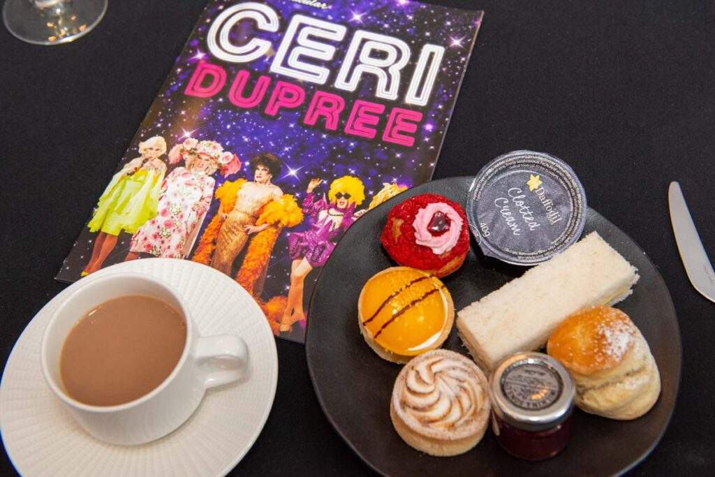 A plate of afternoon tea treats, including sandwiches, cakes and a scone, alongside a cup of tea and a colourful Ceri Dupree souvenir programme.