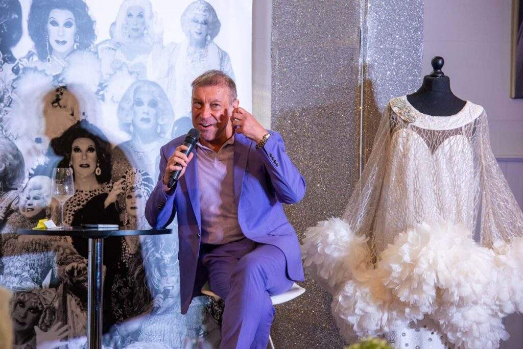 Ceri Dupree speaking into a microphone while gesturing to his head to show where he puts the glue for his wigs. He is wearing a lilac suit in front of a backdrop featuring his glamorous characters.