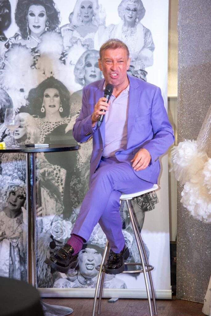 Ceri Dupree sat on his stool and holding his microphone. He is quite animated and pulling a funny face to represent one of his characters. He is clearly mid-story.