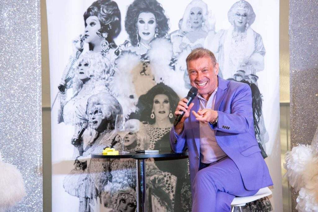 Ceri Dupree smiling and gesturing towards the audience while speaking into a microphone, wearing a lilac suit in front of a backdrop featuring his glamorous characters.