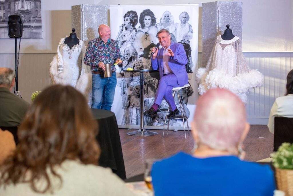 Ceri Dupree laughing on stage alongside another man, with two elaborate white feathered costumes displayed on mannequins either side of them.