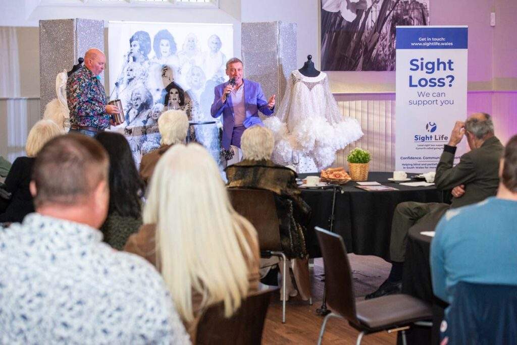 Ceri Dupree entertaining the audience on stage, speaking into a microphone alongside a display of glamorous costumes and a Sight Life banner.