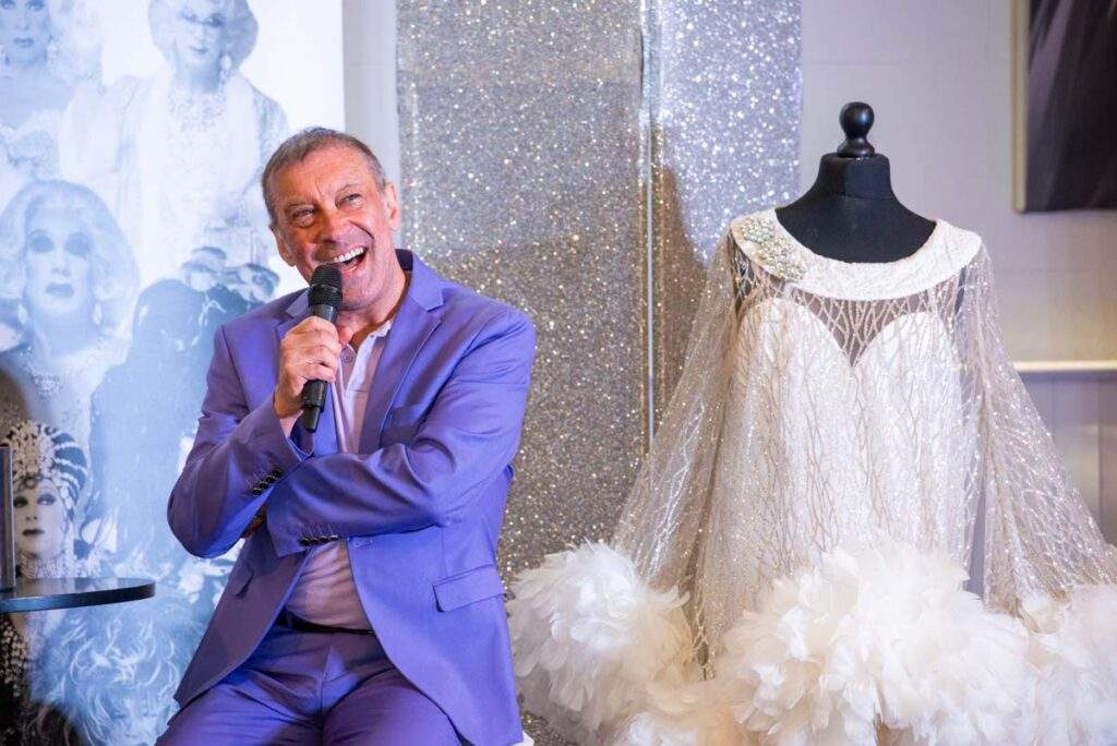 Ceri Dupree laughing into a microphone beside an extravagant white feathered costume, with a sparkling silver backdrop behind him.