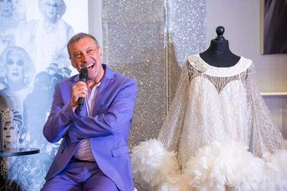 Ceri Dupree laughing into a microphone beside an extravagant white feathered costume, with a sparkling silver backdrop behind him.