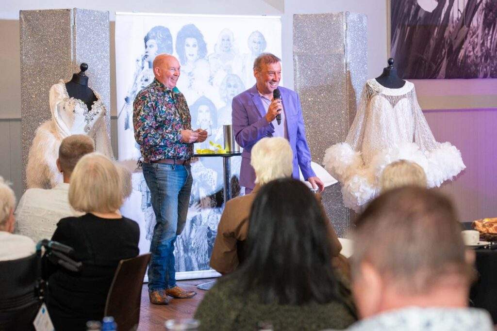 Ceri Dupree laughing on stage alongside another man, with two elaborate white feathered costumes displayed on mannequins either side of them.