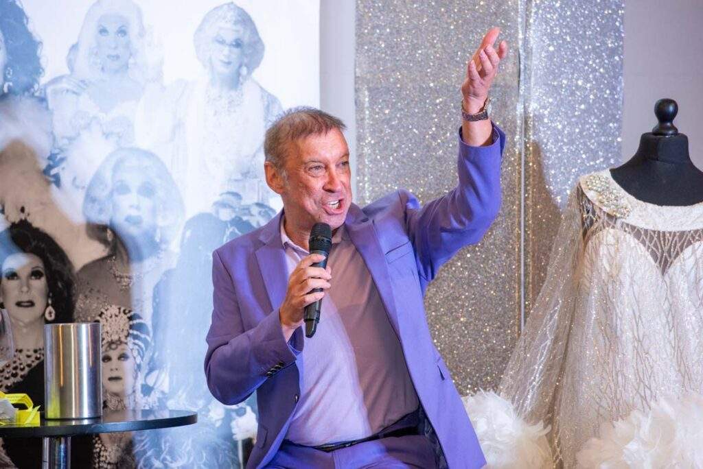 Ceri Dupree enthusiastically addressing the audience, raising one arm while holding a microphone, dressed in a lilac suit beside a glamorous white costume.