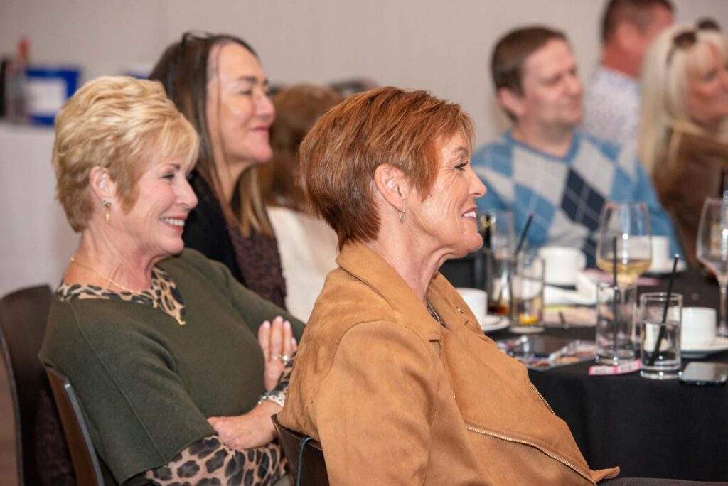 Audience members smiling and enjoying Ceri Dupree's stories, seated around tables with drinks during the afternoon tea event.