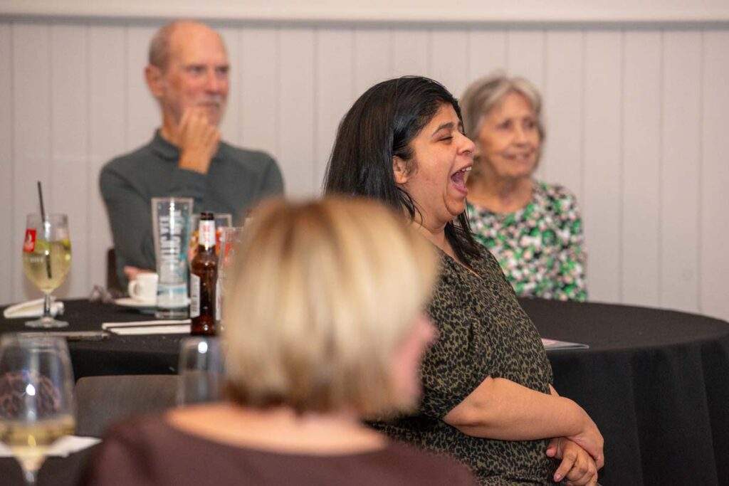 A woman laughing heartily while listening to Ceri Dupree, with other guests seated around her at the event.