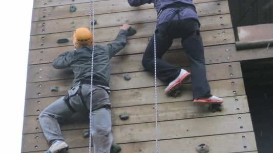 Two people with orange crash helmets climbing a wooden climbing wall with ropes
