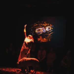 Image of a performer bathed in a red light,with one arm up in the air facing the ceiling, and the other hand holding the lead of her guide dog.