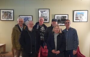 Picture of members of the photography group standing together smiling for the camera, their framed prints on the wall behind them in the Waterloo Tea Rooms.