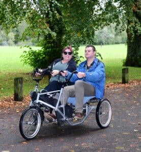 A volunteer and a member cycling on a tandem bike in the park.