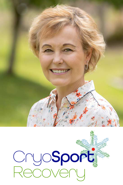 Head and shoulders shot of Andrea standing side on but looking to camera with a big smile. She's outside in a green space with trees and grass out of focus in the background. She's wearing a white blouse with red flowers on it.