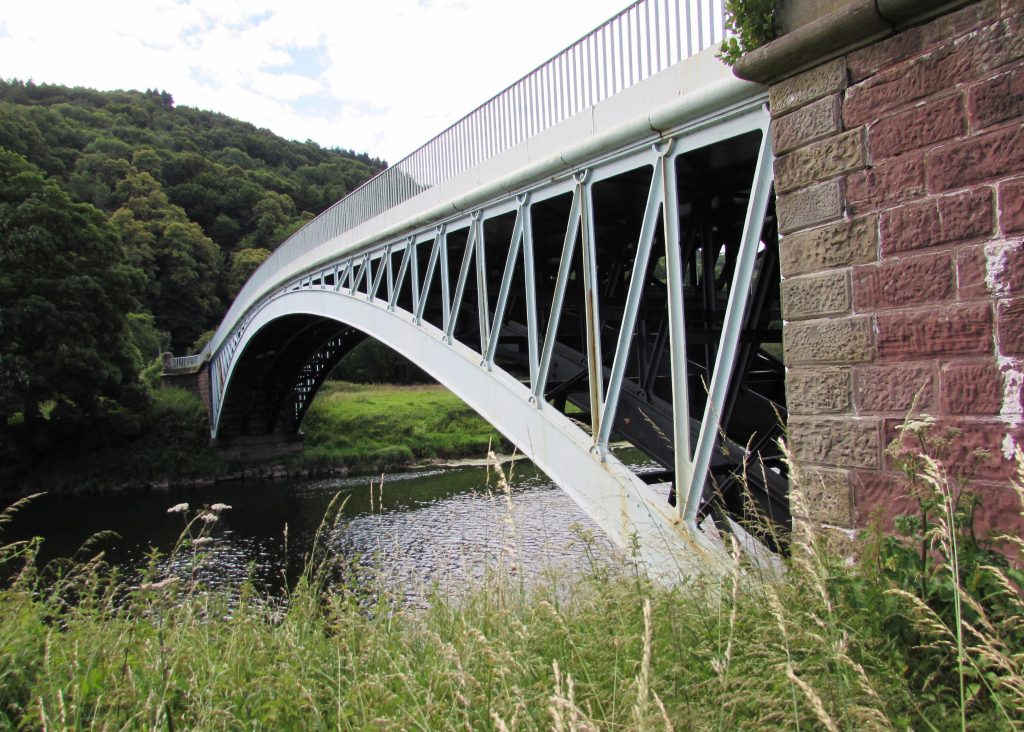 Senedd Photography Exhibition image: A view of a metal bridge over the River Wye taken from close to the bridge entry with the bridge spanning away into the distance. Lots of long grass in the foreground and woodland on the far side.