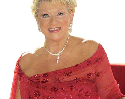 A photo of Beverley sitting and looking to camera in a glamorous red dress and diamante necklace.