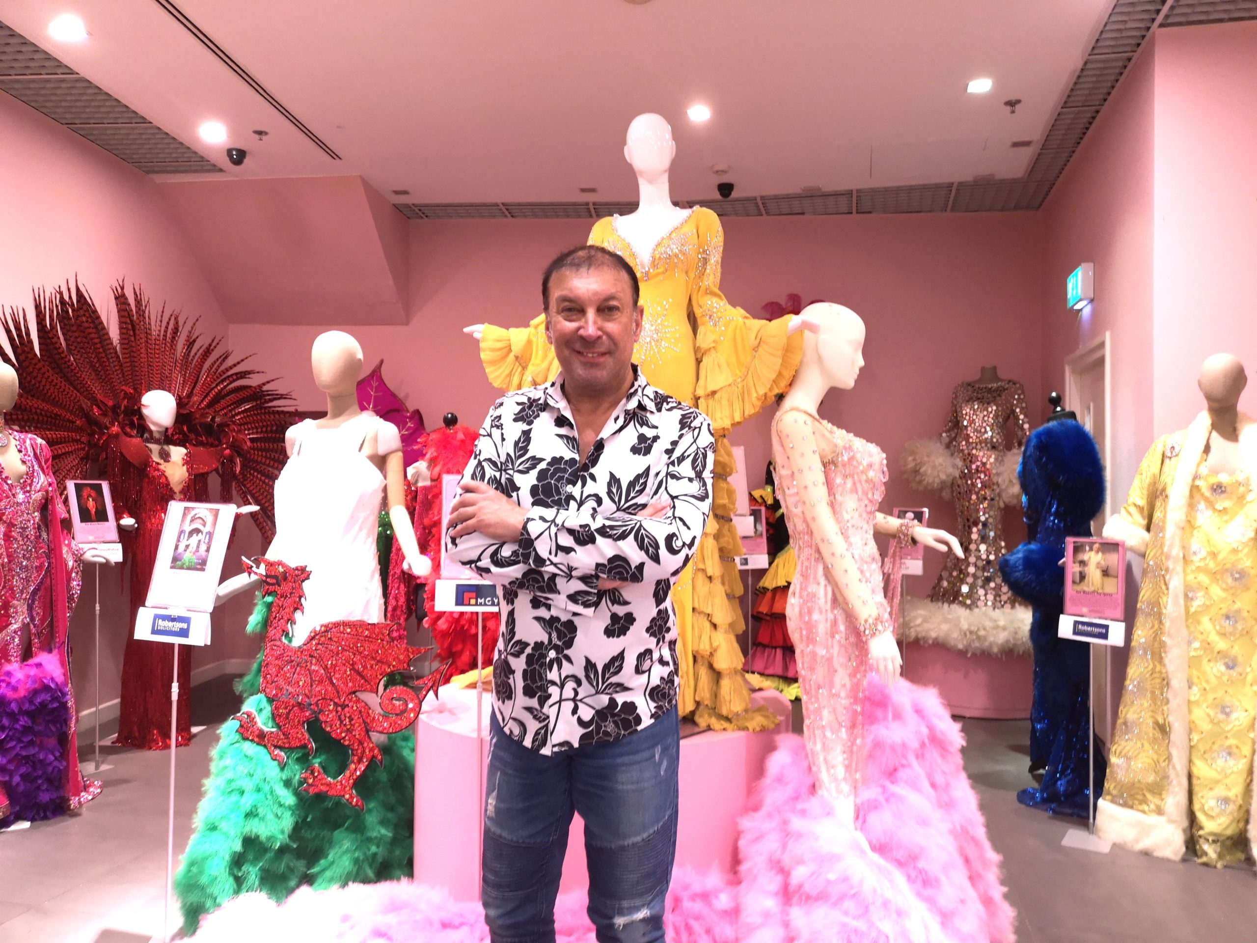 Portrait of Ceri stood with his arms folded in front of several mannequins displaying his gowns.