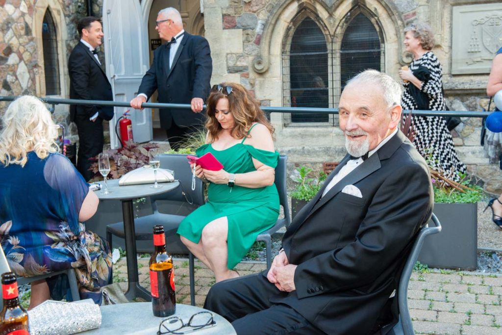 Des Radcliffe smiling to camera. He's sitting outside at a table with a beer. Others are in the background.