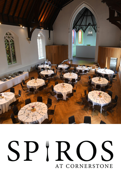 A wide angle image of the beautiful Cornerstone venue's beautiful main hall with vaulted church ceiling and wooden floor. It's laid with 12+ round tables for an event. There's a stage area at the back of the room. Under the image is the Spiros at Cornerstone logo