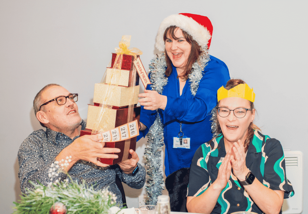 Three people enjoying a festive celebration at a Sight Life Christmas party. A woman wearing a Santa hat and silver tinsel presents a stack of wrapped raffle prizes to a smiling man, while a woman wearing a yellow paper Christmas crown claps excitedly.