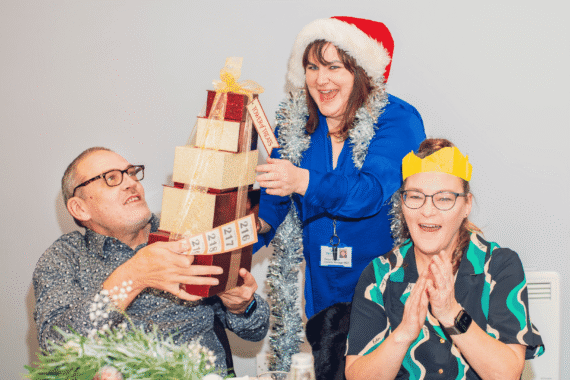 Three people enjoying a festive celebration at a Sight Life Christmas party. A woman wearing a Santa hat and silver tinsel presents a stack of wrapped raffle prizes to a smiling man, while a woman wearing a yellow paper Christmas crown claps excitedly.
