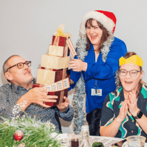 Three people enjoying a festive celebration at a Sight Life Christmas party. A woman wearing a Santa hat and silver tinsel presents a stack of wrapped raffle prizes to a smiling man, while a woman wearing a yellow paper Christmas crown claps excitedly.